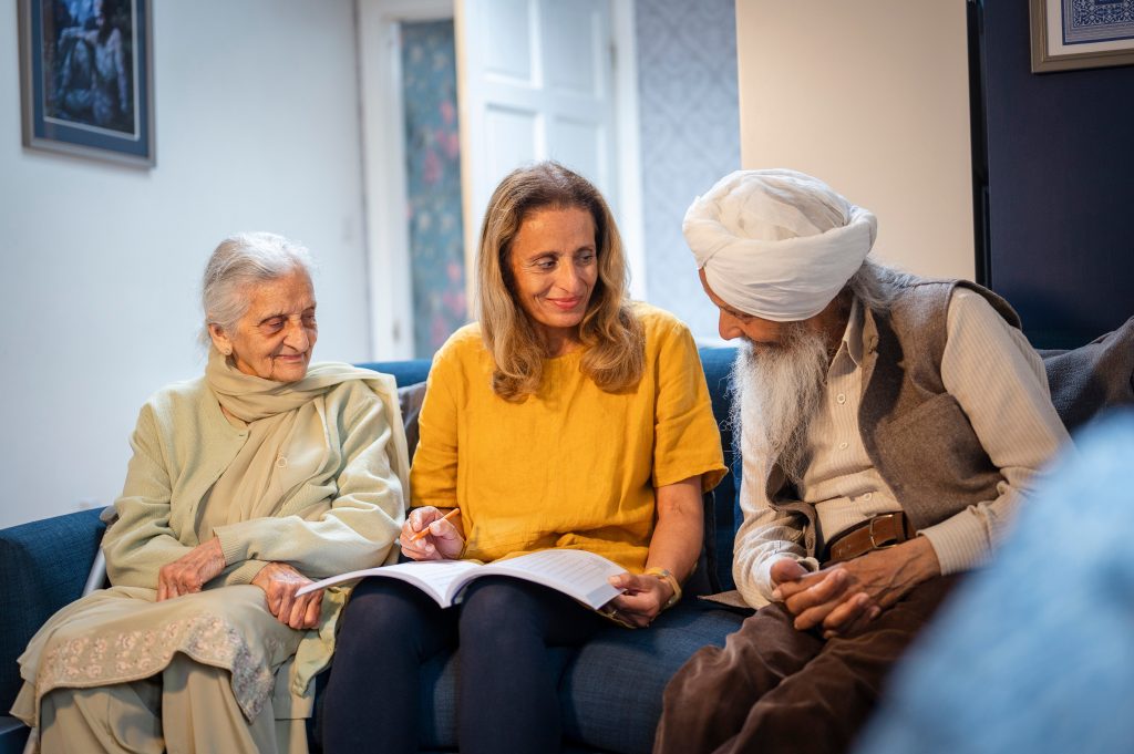 Two older adult parents sit on a couch with their adult daughter between them, looking at a book in her lap