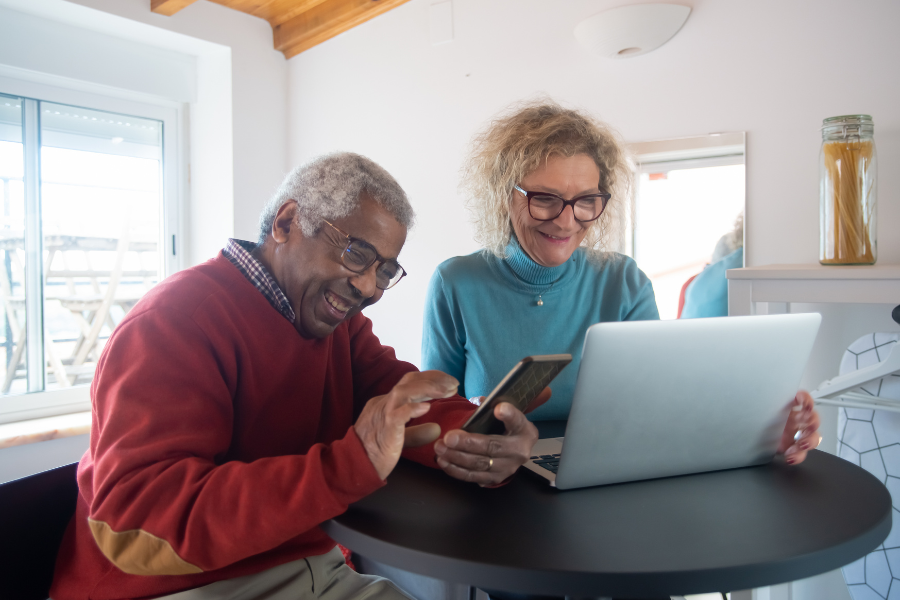 An older man laughs at something on his phone, while an older woman smiles next to him looking at a laptop screen