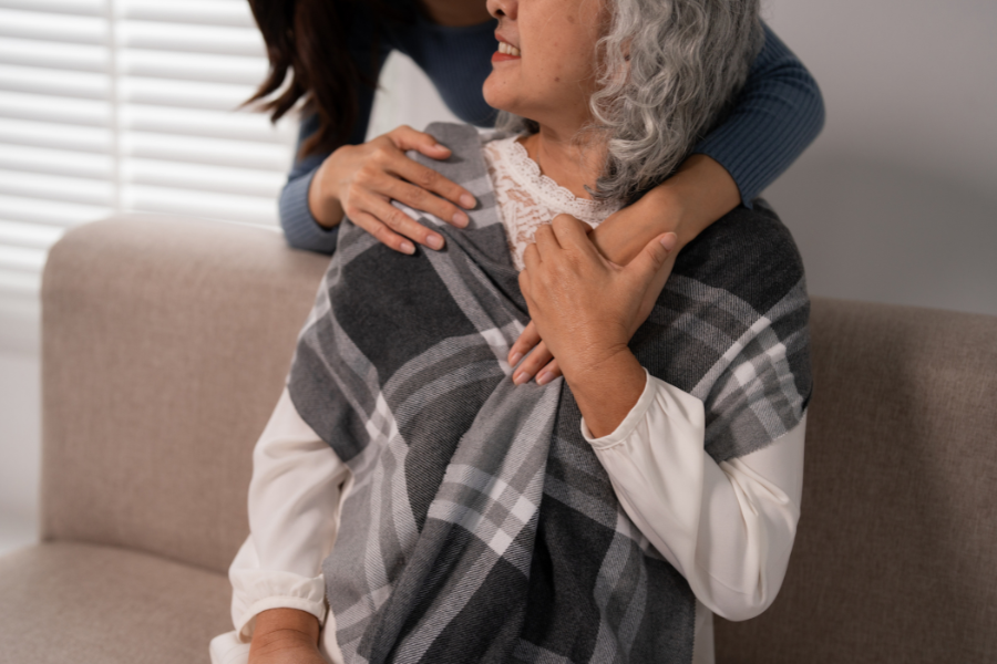 An older adult sat on the couch turns as another person hugs them from behind